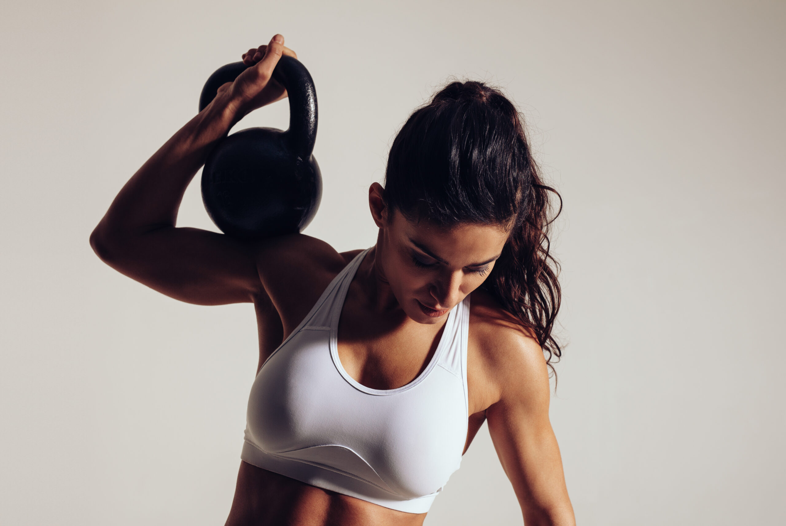 Determined young fitness woman exercising with kettle bell on grey background. Woman in sportswear doing crossfit workout with one hand.